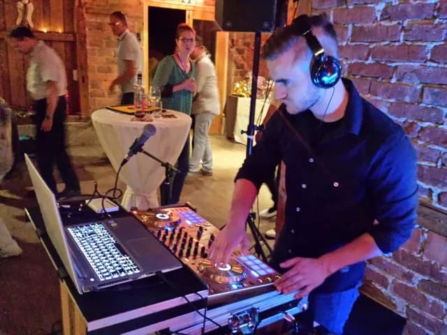 DJ mit Kopfhörern legt an seinem Pult in einer rustikalen Scheune mit Backsteinwand auf, im Hintergrund Hochzeitsgäste